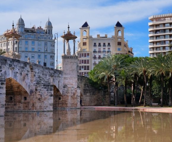 A scenic view of a bridge with ornate arches, surrounded by palm trees and historic buildings reflecting in a calm body of water under a blue sky.
