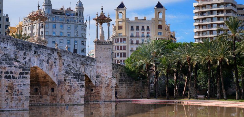 A scenic view of a bridge with ornate arches, surrounded by palm trees and historic buildings reflecting in a calm body of water under a blue sky.