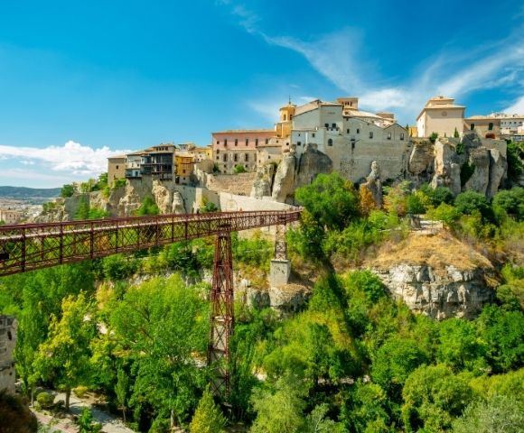 A picturesque view of a rocky landscape featuring a historic village on a hill, with a bridge connecting the land across a green valley under a bright blue sky.