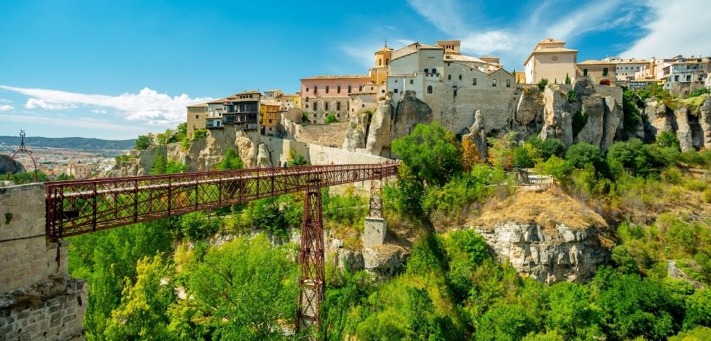 A picturesque view of a rocky landscape featuring a historic village on a hill, with a bridge connecting the land across a green valley under a bright blue sky.