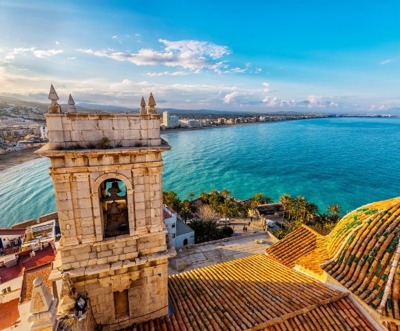 A scenic view of a coastline featuring a bell tower and lush greenery, with buildings along the shore under a blue sky.