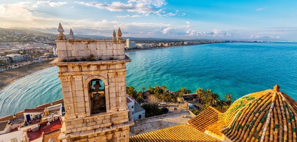 A scenic view of a coastline featuring a bell tower and lush greenery, with buildings along the shore under a blue sky.