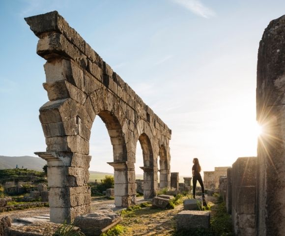 A person standing near ancient stone ruins with arches, illuminated by the setting sun.