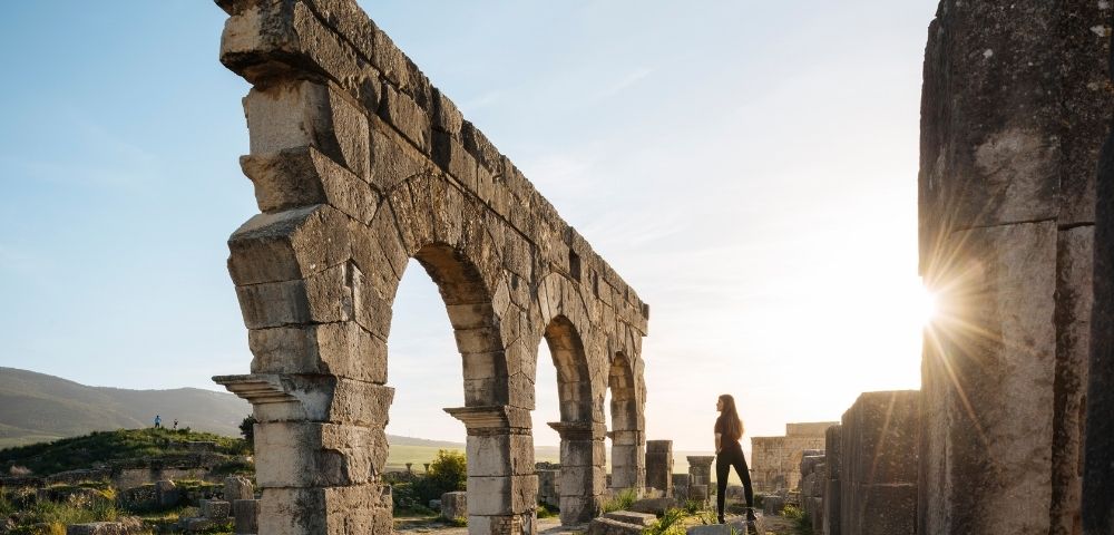 A person standing near ancient stone ruins with arches, illuminated by the setting sun.
