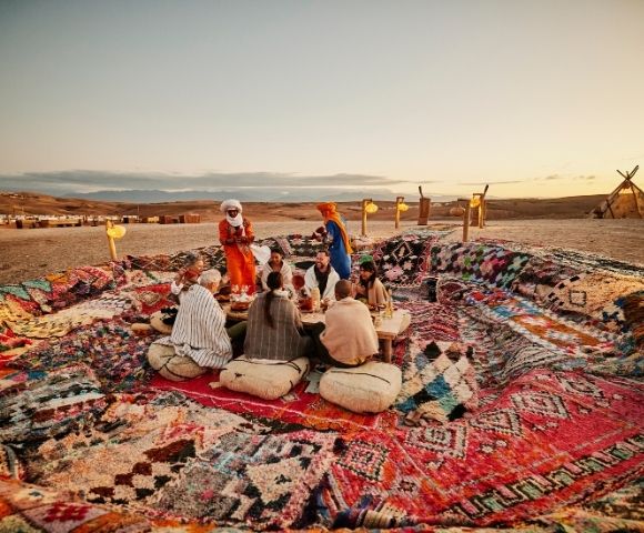 A group of people sitting on a colorful rug in a desert setting, engaging in conversation as the sun sets.