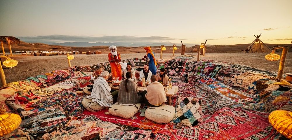 A group of people sitting on a colorful rug in a desert setting, engaging in conversation as the sun sets.