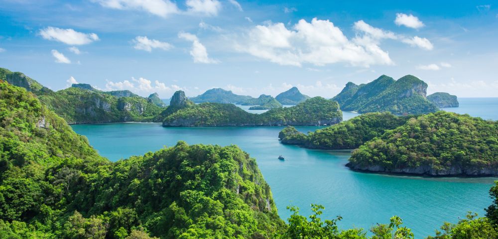 A panoramic view of numerous green, jungle-covered islands with steep rocky cliffs rising from turquoise waters under a bright blue sky with fluffy white clouds.