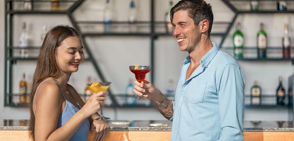 Smiling couple raise colorful cocktails at a stylish bar, with liquor bottles on geometric shelves; warm, relaxed, sociable atmosphere.