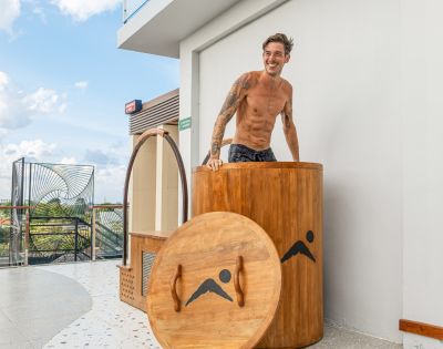 Smiling tattooed shirtless man stands behind a wooden barrel sauna beside its round lid on a sunny modern patio with tropical greenery.