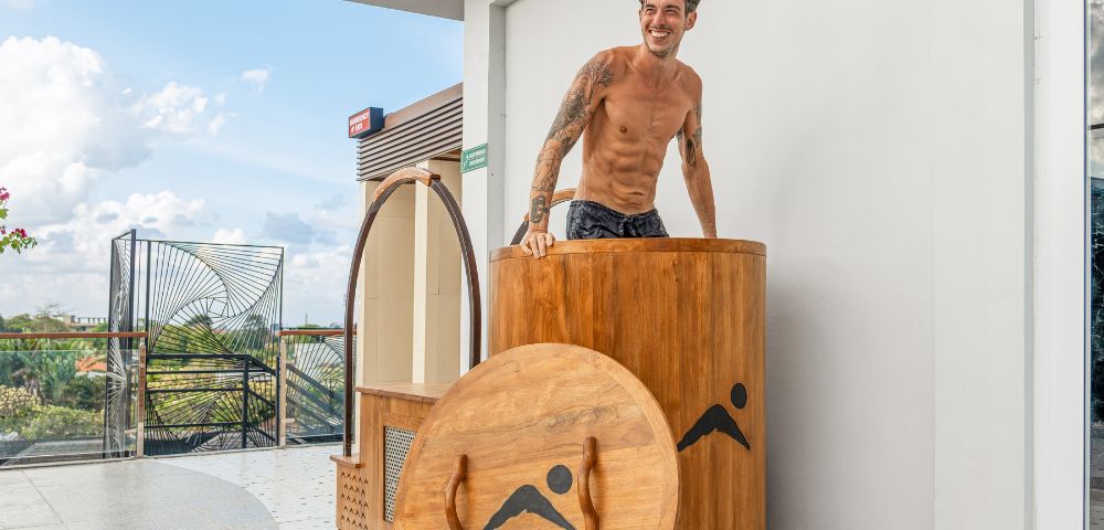 Smiling tattooed shirtless man stands behind a wooden barrel sauna beside its round lid on a sunny modern patio with tropical greenery.