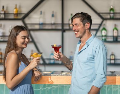 Smiling couple raise colorful cocktails at a stylish bar, with liquor bottles on geometric shelves; warm, relaxed, sociable atmosphere.