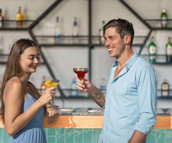 Smiling couple raise colorful cocktails at a stylish bar, with liquor bottles on geometric shelves; warm, relaxed, sociable atmosphere.