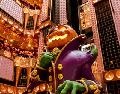 A spooky, illuminated jack-o'-lantern character with green hands and a purple coat adorned with skulls stands in a richly decorated atrium with warm lighting.
