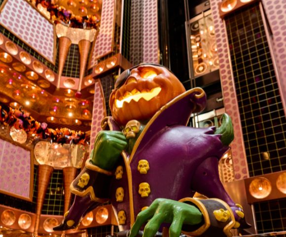 A spooky, illuminated jack-o'-lantern character with green hands and a purple coat adorned with skulls stands in a richly decorated atrium with warm lighting.