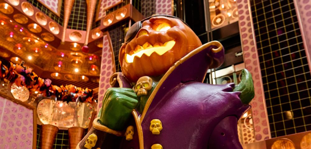 A spooky, illuminated jack-o'-lantern character with green hands and a purple coat adorned with skulls stands in a richly decorated atrium with warm lighting.