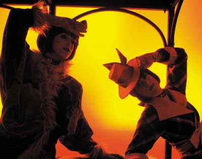 Two figures in elaborate costumes are illuminated against a vibrant yellow background, suggesting a theatrical performance. The figure on the left wears a furry, shaggy costume and a dark cap, with one hand raised to their forehead. The figure on the right is dressed in a checkered garment and a distinctive orange hat with pointed ear-like accents, their head tilted down.