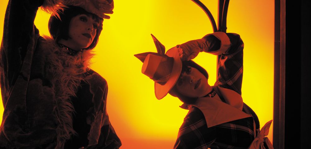Two figures in elaborate costumes are illuminated against a vibrant yellow background, suggesting a theatrical performance. The figure on the left wears a furry, shaggy costume and a dark cap, with one hand raised to their forehead. The figure on the right is dressed in a checkered garment and a distinctive orange hat with pointed ear-like accents, their head tilted down.
