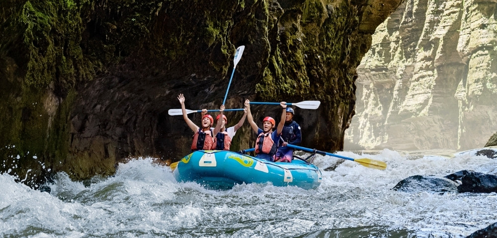 Four rafters paddle a blue inflatable boat through foaming whitewater beneath a mossy canyon arch, conveying adventure and excitement.