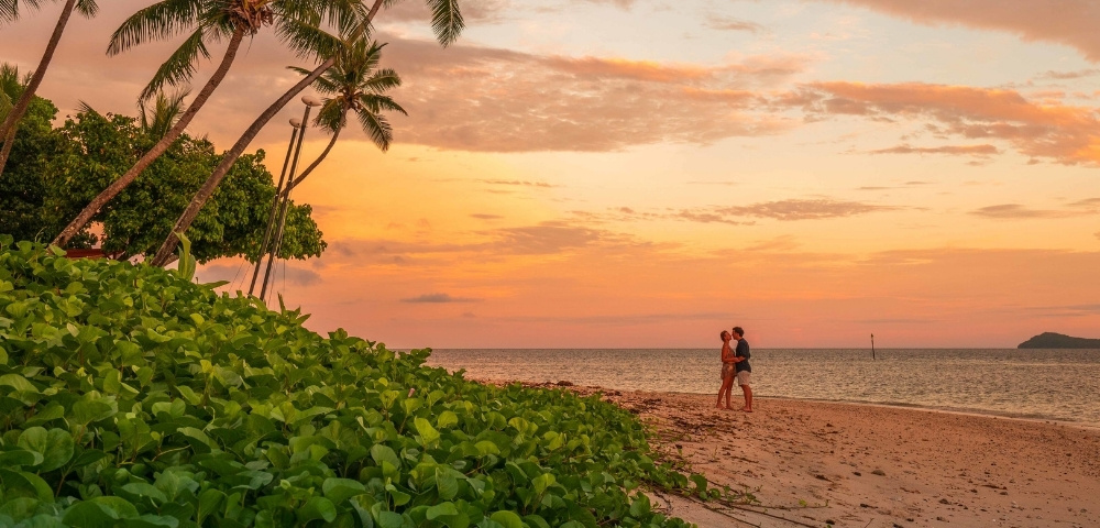 Tropical beach at sunset with leaning palm trees, lush greenery, and a couple strolling beside the calm ocean in warm orange light.