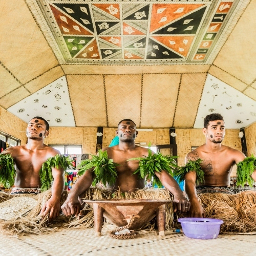 Three men in leafy garlands and woven skirts sit in a traditional hut, beneath a colorful geometric ceiling, creating a vibrant cultural scene.