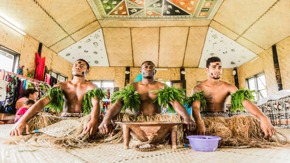 Three men in leafy garlands and woven skirts sit in a traditional hut, beneath a colorful geometric ceiling, creating a vibrant cultural scene.