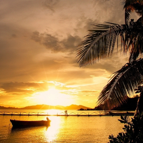 Golden sunset over calm tropical water, with palm fronds, a silhouetted boat, distant mountains, and two people on a pier; tranquil mood.