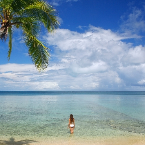 Palm-fringed tropical beach with a woman wading into calm, clear turquoise water beneath a bright blue, cloud-filled sky.