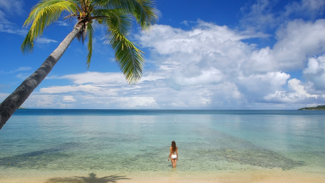 Palm-fringed tropical beach with a woman wading into calm, clear turquoise water beneath a bright blue, cloud-filled sky.