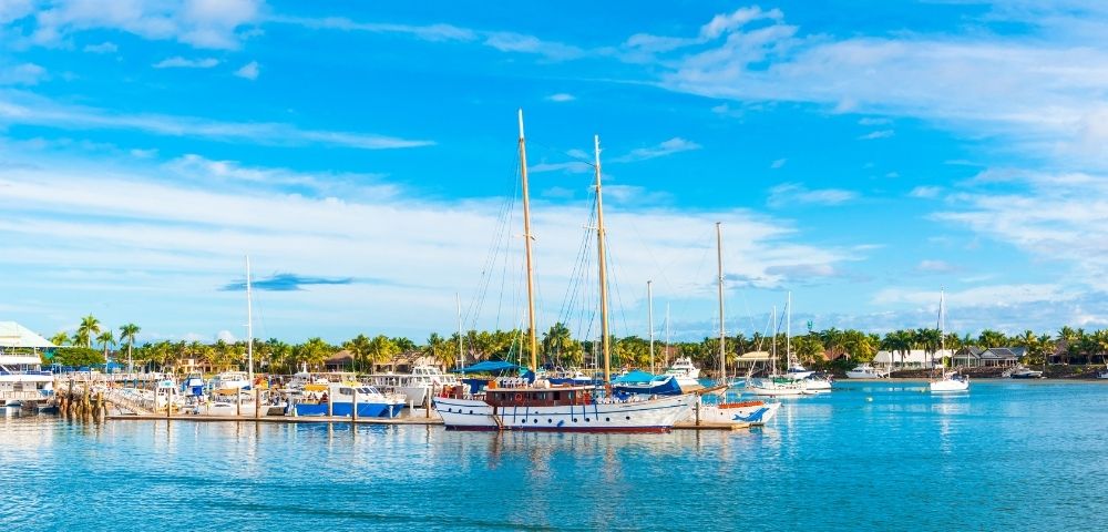Bright blue tropical marina with sailboats and yachts moored on calm turquoise water, palm trees along shore beneath a cloud-streaked sky.
