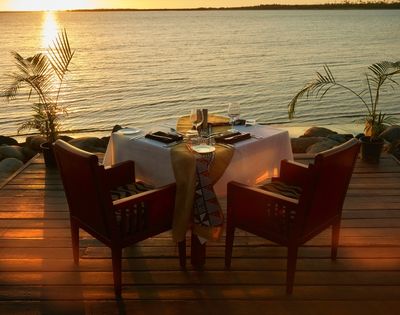 Sunset waterfront dinner table on a wooden deck, set for two with candles and red chairs, glowing over calm water; romantic, serene mood.