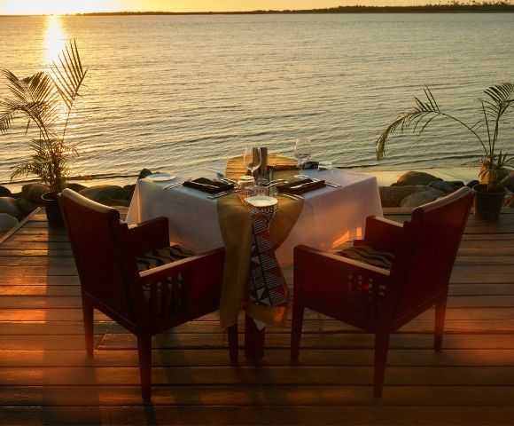 Sunset waterfront dinner table on a wooden deck, set for two with candles and red chairs, glowing over calm water; romantic, serene mood.