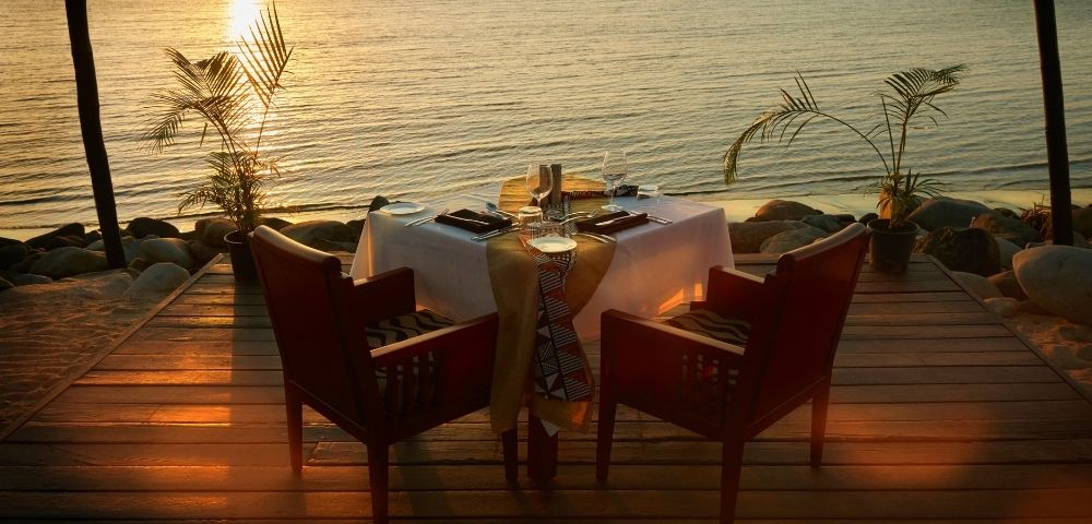 Sunset waterfront dinner table on a wooden deck, set for two with candles and red chairs, glowing over calm water; romantic, serene mood.