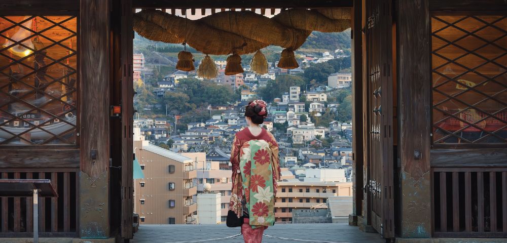 A person in a vibrant floral kimono stands framed by an ancient wooden gateway, looking out at a sprawling Japanese town nestled on a hillside.