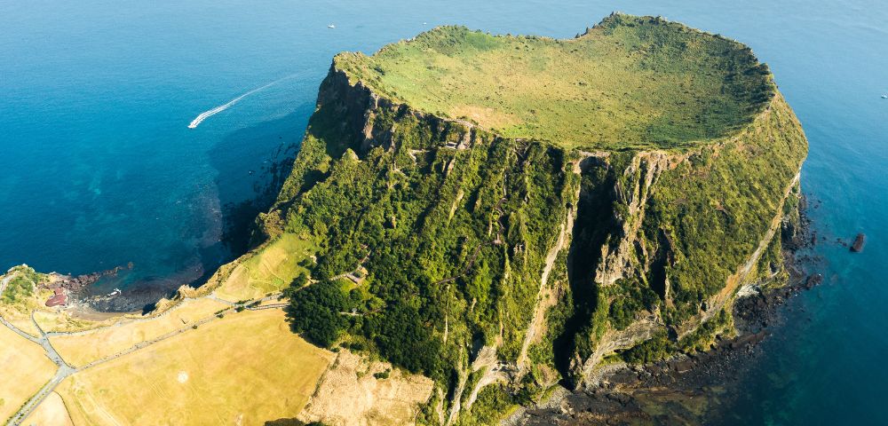 Aerial view of a lush green volcanic crater island with steep cliffs surrounded by blue ocean, and a small boat in the distance.