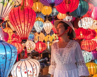 A woman in a white dress and traditional hat stands among colorful hanging lanterns glowing in warm light.
