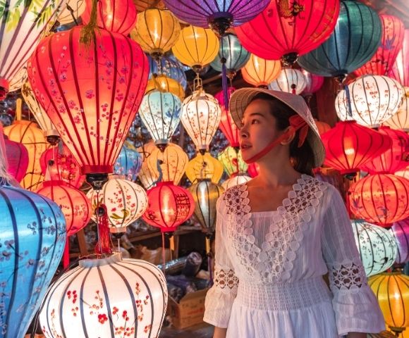 A woman in a white dress and traditional hat stands among colorful hanging lanterns glowing in warm light.