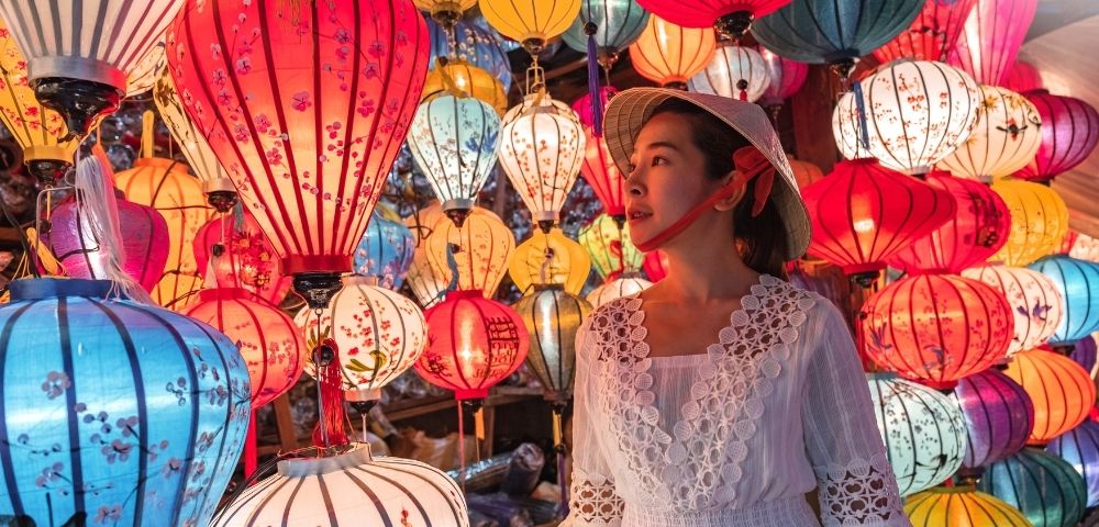 A woman in a white dress and traditional hat stands among colorful hanging lanterns glowing in warm light.