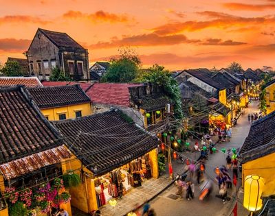 Sunset over a bustling street in a historic town with yellow buildings, tiled roofs, and colorful lanterns, filled with moving pedestrians.
