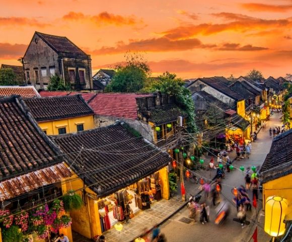 Sunset over a bustling street in a historic town with yellow buildings, tiled roofs, and colorful lanterns, filled with moving pedestrians.