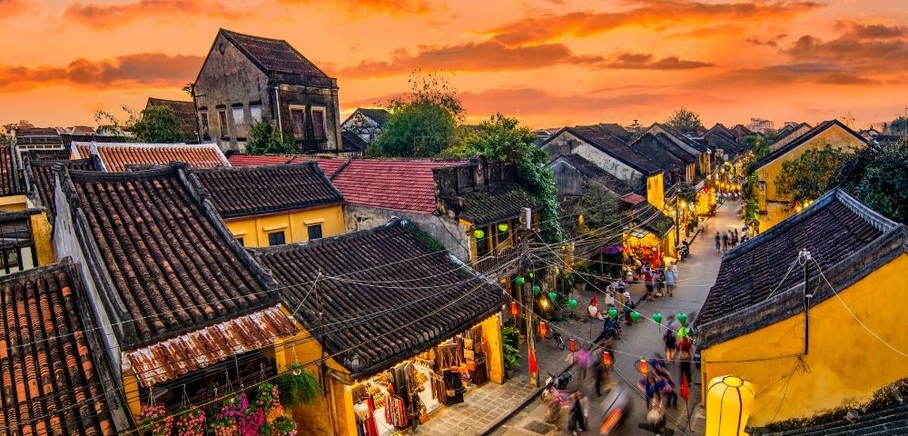 Sunset over a bustling street in a historic town with yellow buildings, tiled roofs, and colorful lanterns, filled with moving pedestrians.