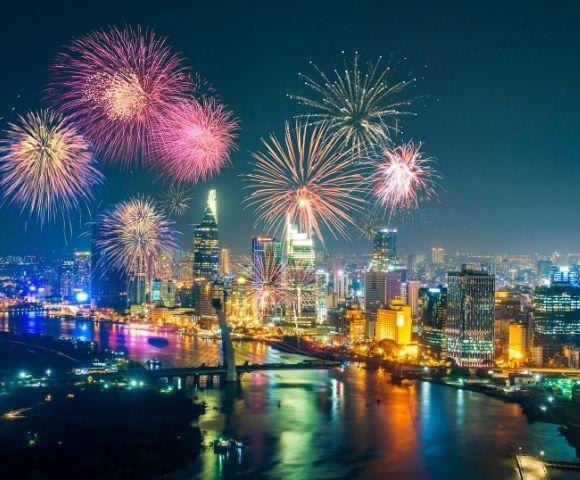 Nighttime cityscape with colorful fireworks bursting over illuminated skyscrapers and a river reflecting vibrant lights.