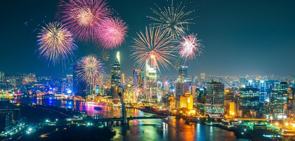 Nighttime cityscape with colorful fireworks bursting over illuminated skyscrapers and a river reflecting vibrant lights.