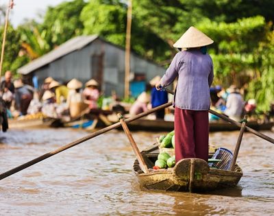 Person wearing a conical hat rowing a wooden boat filled with produce on a river at a bustling floating market.