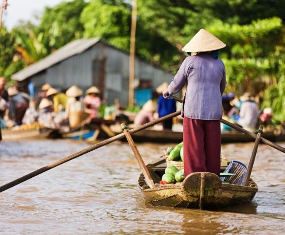 Person wearing a conical hat rowing a wooden boat filled with produce on a river at a bustling floating market.
