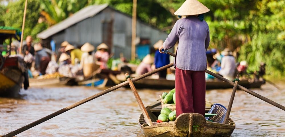 Person wearing a conical hat rowing a wooden boat filled with produce on a river at a bustling floating market.