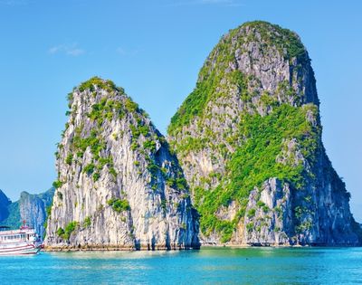 Boat sailing near two large, green-covered limestone karst formations rising from clear blue water under a bright sky.