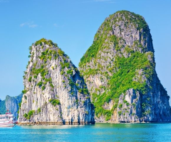 Boat sailing near two large, green-covered limestone karst formations rising from clear blue water under a bright sky.