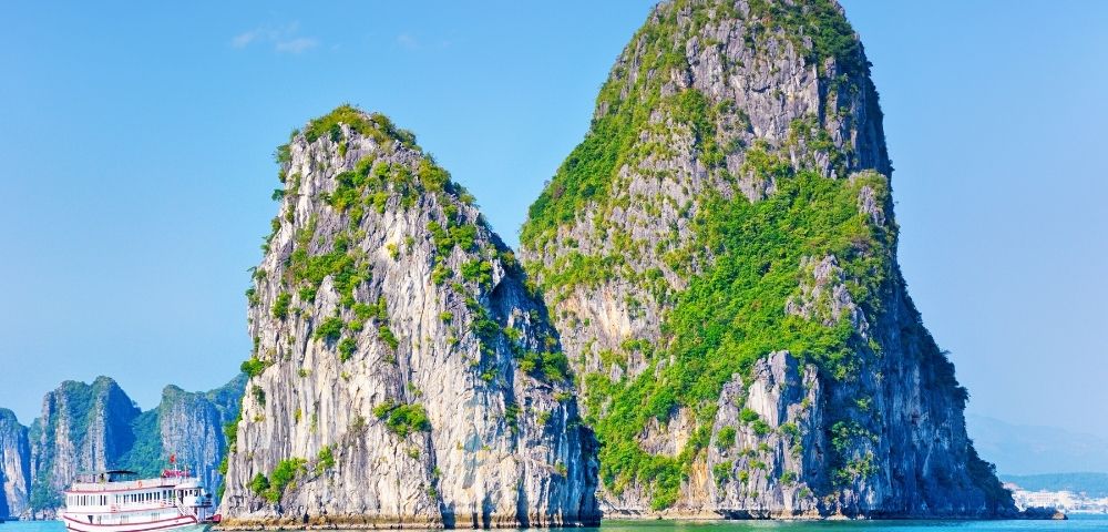 Boat sailing near two large, green-covered limestone karst formations rising from clear blue water under a bright sky.