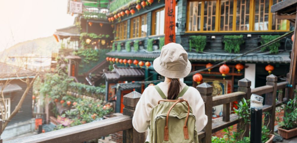 A person wearing a hat and backpack stands on a wooden balcony, looking out at traditional buildings adorned with red lanterns and lush greenery.