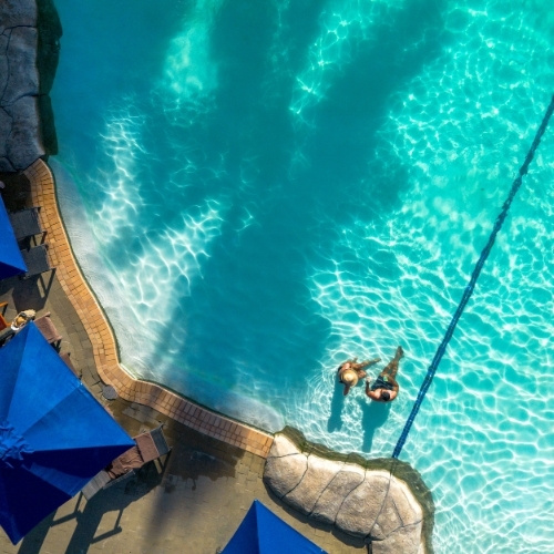 Aerial view of two swimmers sitting in a bright turquoise pool beside a curved stone deck, with sunlit ripples and blue umbrellas.